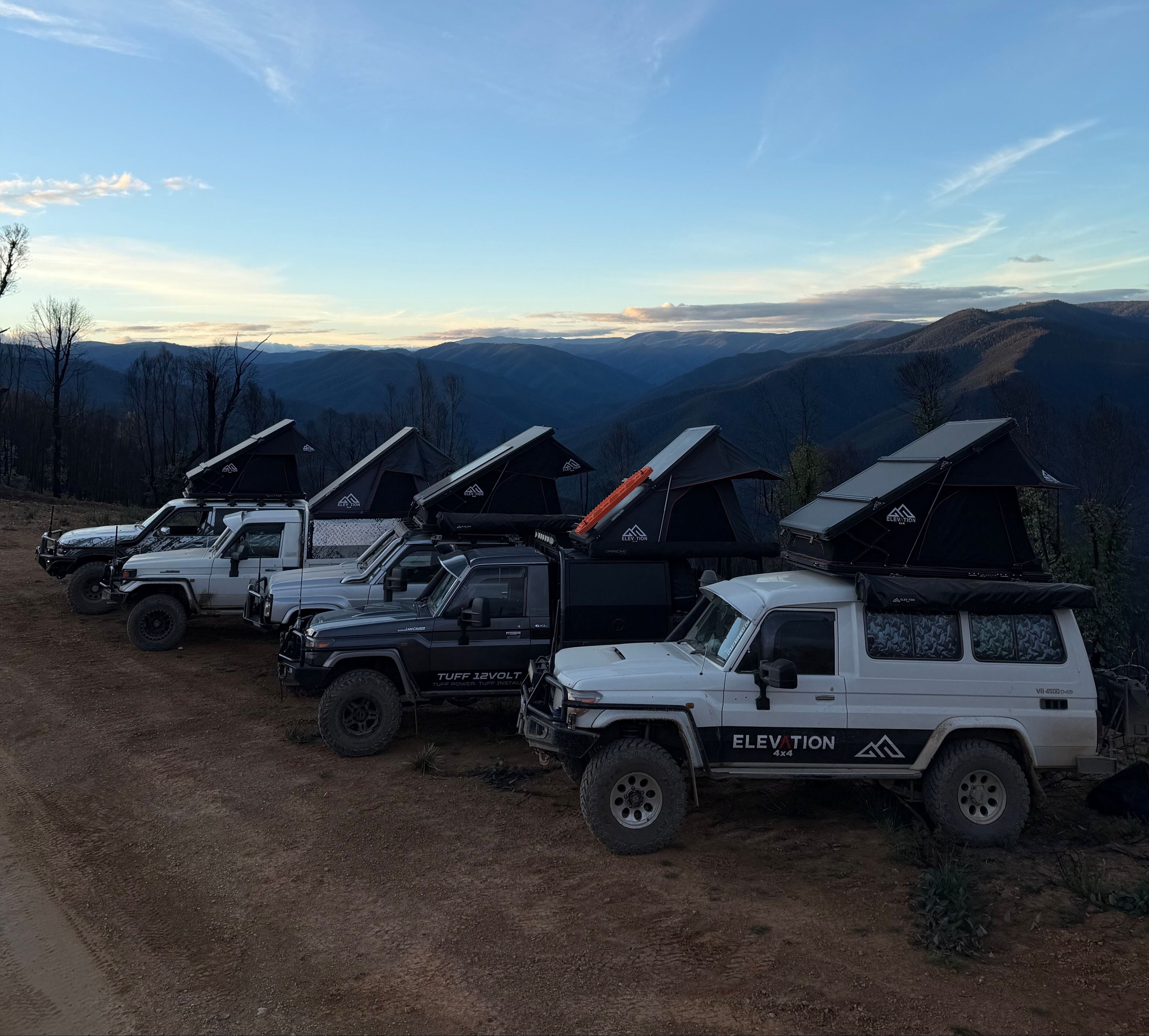 Row of off-road vehicles with rooftop tents parked on a dirt road with mountains in the background.